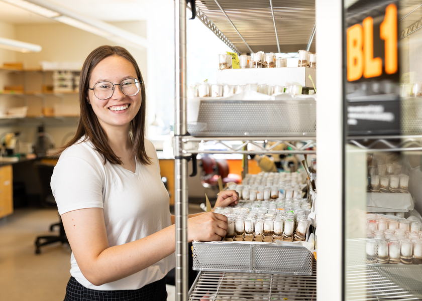 A smiling researcher in a white t-shirt stands beside an open laboratory refrigerator stocked with rows of small sample vials.