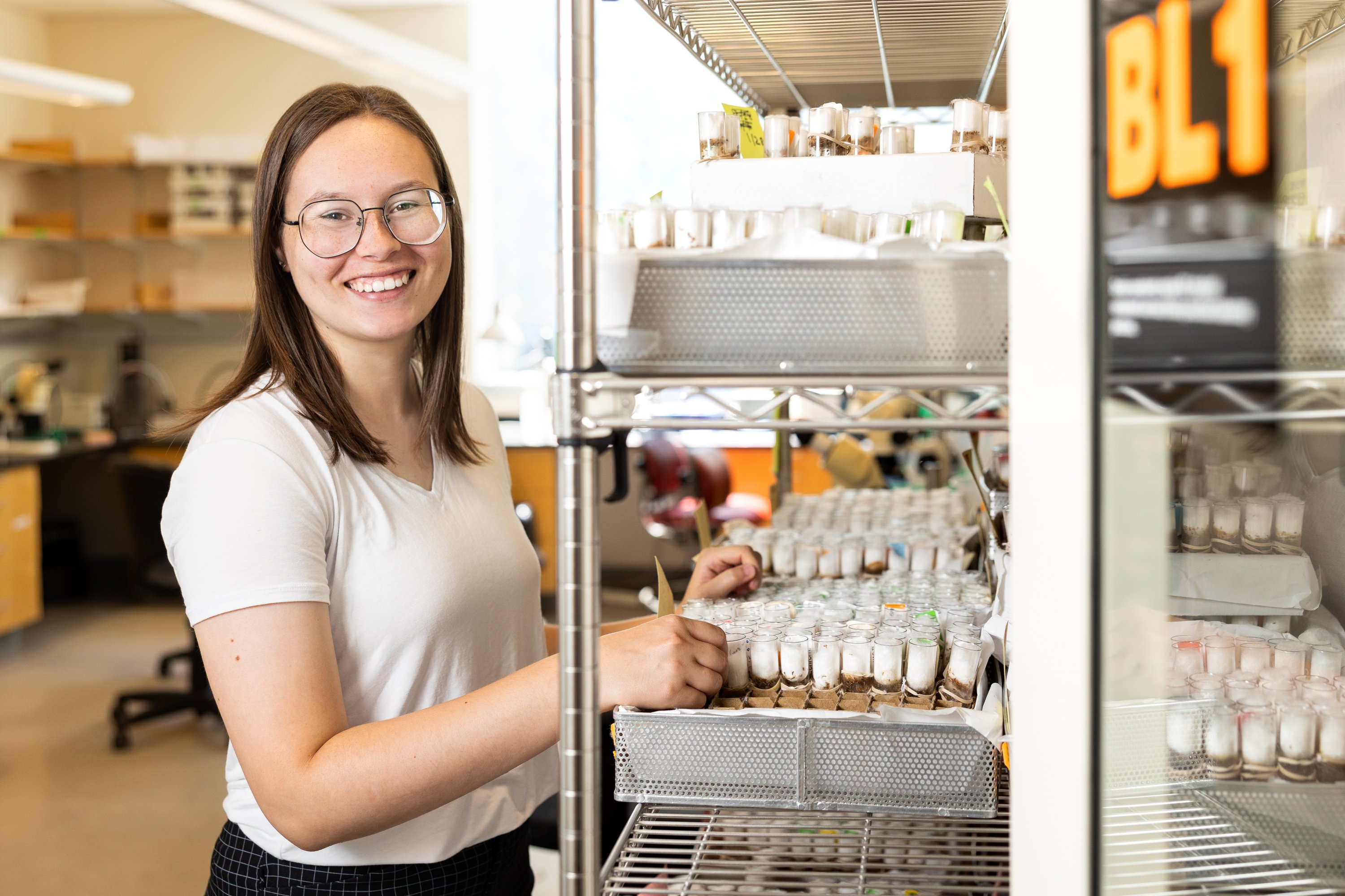 A smiling researcher in a white t-shirt stands beside an open laboratory refrigerator stocked with rows of small sample vials.