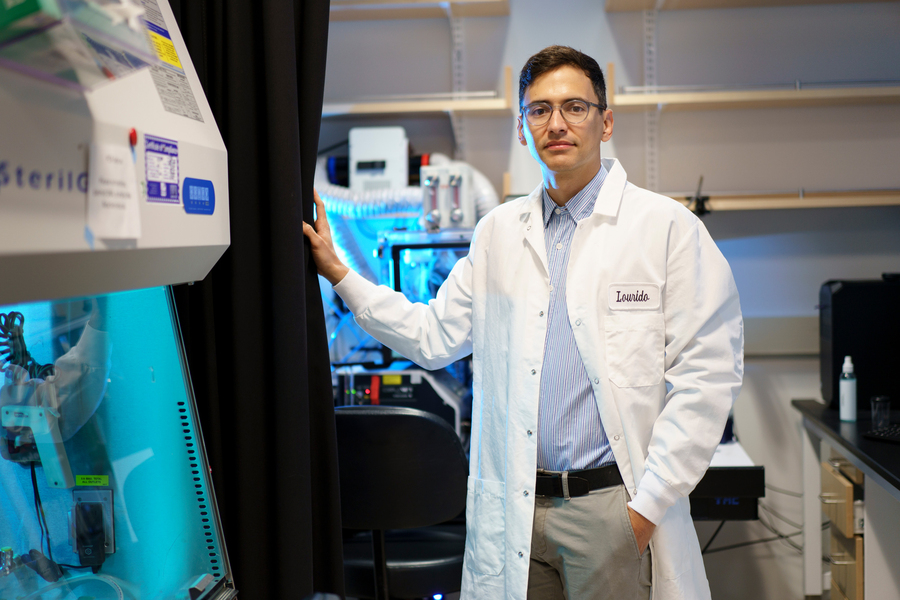 A researcher in a white lab coat labeled "Lourido" stands beside a biosafety cabinet in a blue-lit laboratory.