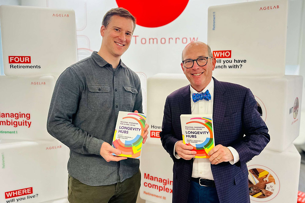 Two men stand side by side holding copies of a book titled "Longevity Hubs" in front of an MIT AgeLab branded display.