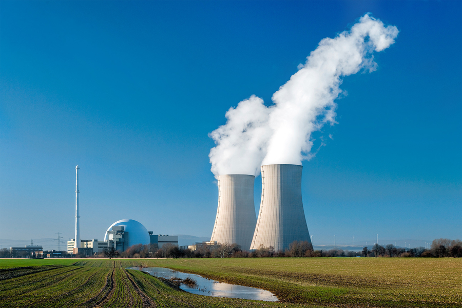 Three large cooling towers emit steam against a clear blue sky at a nuclear power plant, with a reactor dome and open farmland visible in the foreground.
