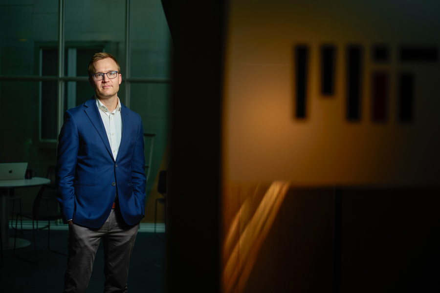 A man in a blue blazer and glasses stands in a dimly lit corridor with a blurred MIT sign visible in the warm-toned background.