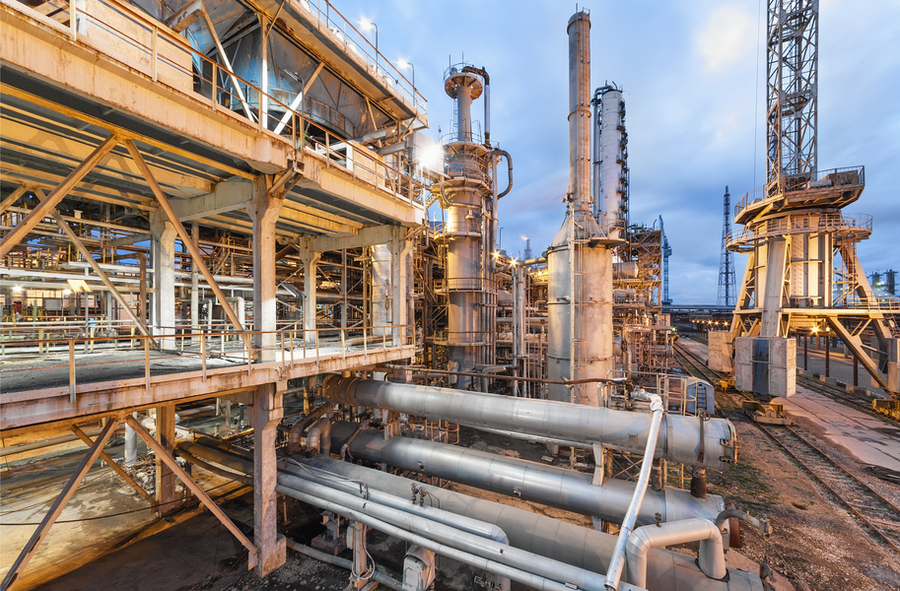 An industrial refinery at dusk with large pipes, steel frameworks, distillation towers, and cranes illuminated against a cloudy sky.
