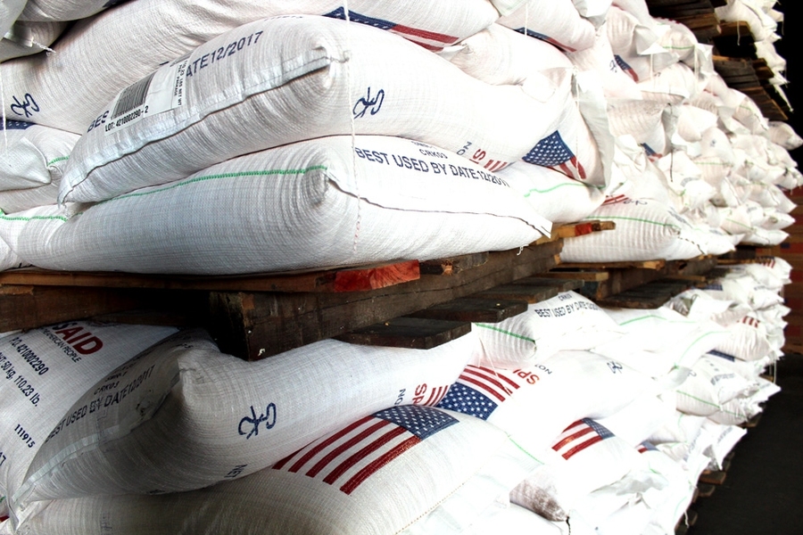 Rows of white USAID food aid sacks stamped with American flags and best-by dates stacked on wooden pallets in a warehouse