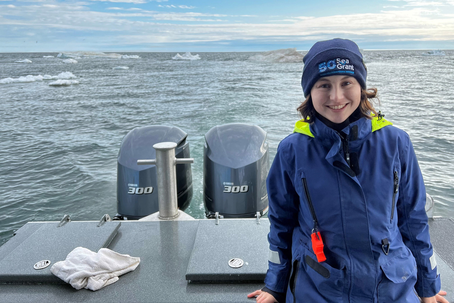 A smiling MIT Sea Grant researcher in a navy jacket and beanie stands on the stern of a research vessel with twin 300-horsepower outboard motors, with icebergs and open Arctic water visible in the background.