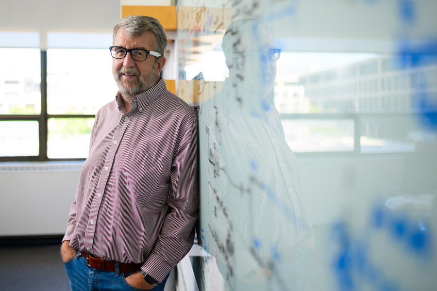 A man with gray hair and glasses leans against a whiteboard covered in equations and diagrams inside a bright office.