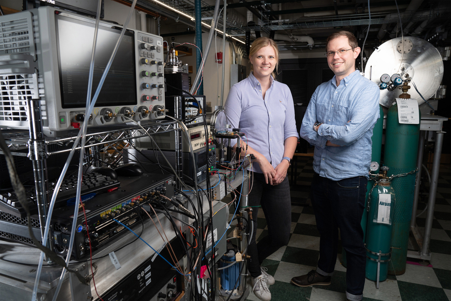 Two MIT researchers stand together in a laboratory filled with rack-mounted electronics, oscilloscopes, and Airgas cylinders, smiling for a portrait related to space carbon detection research.