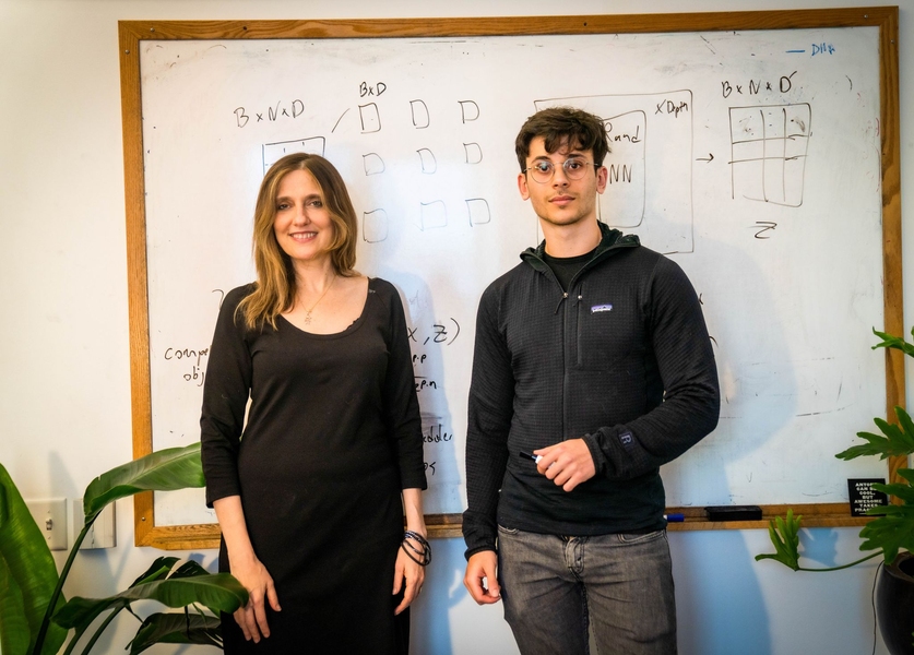 Two MIT researchers standing before a whiteboard covered in machine learning diagrams and matrix notation.