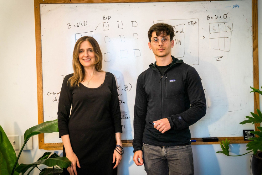 Two MIT researchers standing before a whiteboard covered in machine learning diagrams and matrix notation.