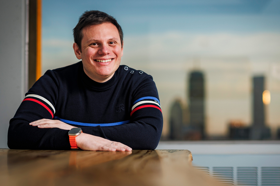 A man in a navy striped sweater leans on a wooden table smiling, with a city skyline glowing at sunset visible through a large window behind him