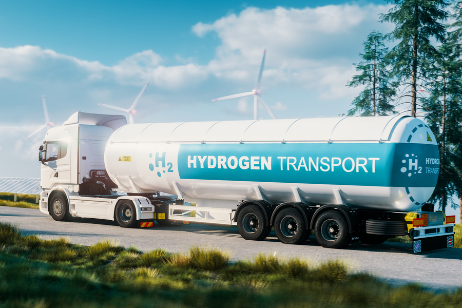A semi-truck towing a large tank labeled "H2 Hydrogen Transport" past wind turbines and solar panels.