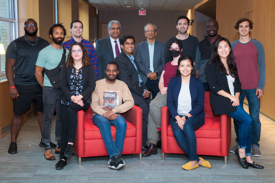 A diverse group of about a dozen researchers poses together in a bright hallway, with some seated in red chairs and others standing behind them.