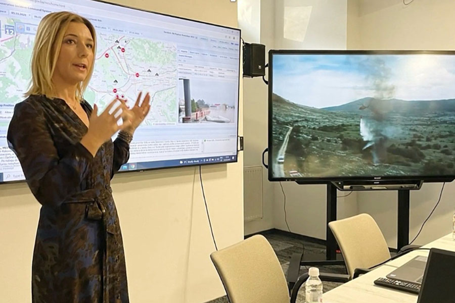 A presenter in a patterned dress gestures toward two large screens displaying an emergency management mapping interface and a wildfire aerial footage in a conference room.