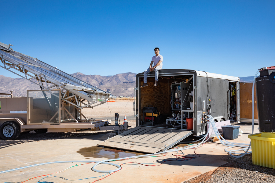 A researcher sits atop a utility trailer at an arid desert field test site beside angled solar collector panels, with dry mountains and clear blue sky in the background.