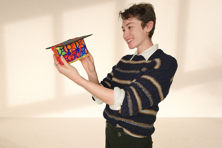 A young MIT student in a striped sweater holds up and admires a small colorful 3D-printed geometric model shaped like a mortarboard cap against a cream-colored wall.