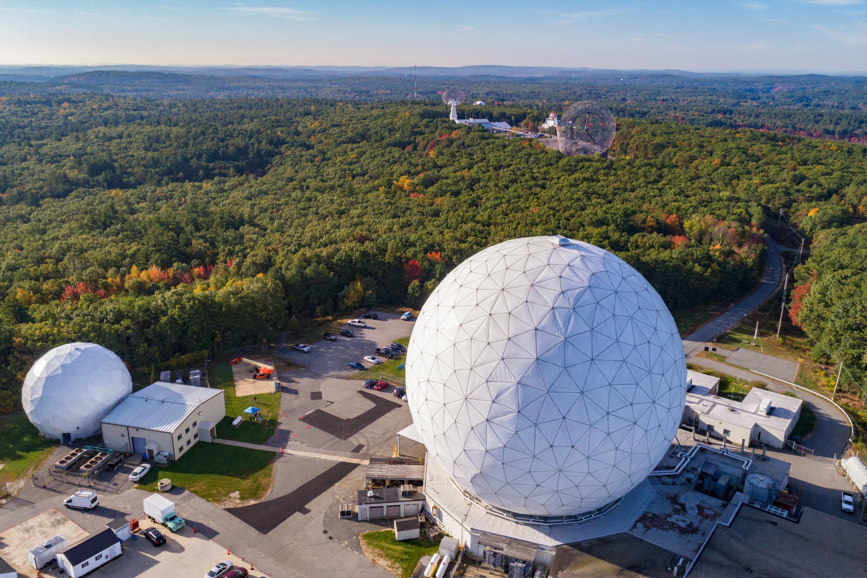Aerial view of a research facility with two large white geodesic radome structures surrounded by fall foliage and wooded landscape.