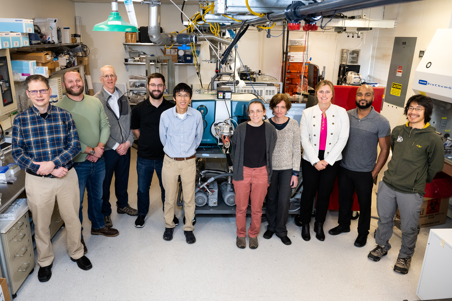 A research team of ten people poses in front of large laboratory equipment inside a well-equipped research facility.