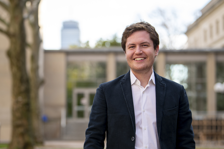A smiling MIT researcher in a dark blazer stands outdoors on a campus pathway with trees and academic buildings softly blurred in the background.