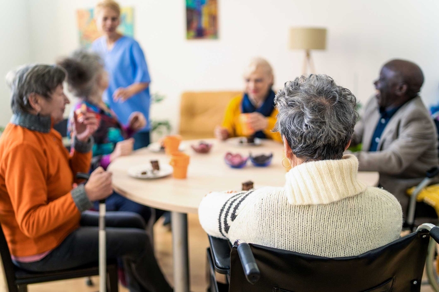 A diverse group of older adults sits around a round table in a bright common room, sharing snacks and conversation while a caregiver in scrubs stands nearby.