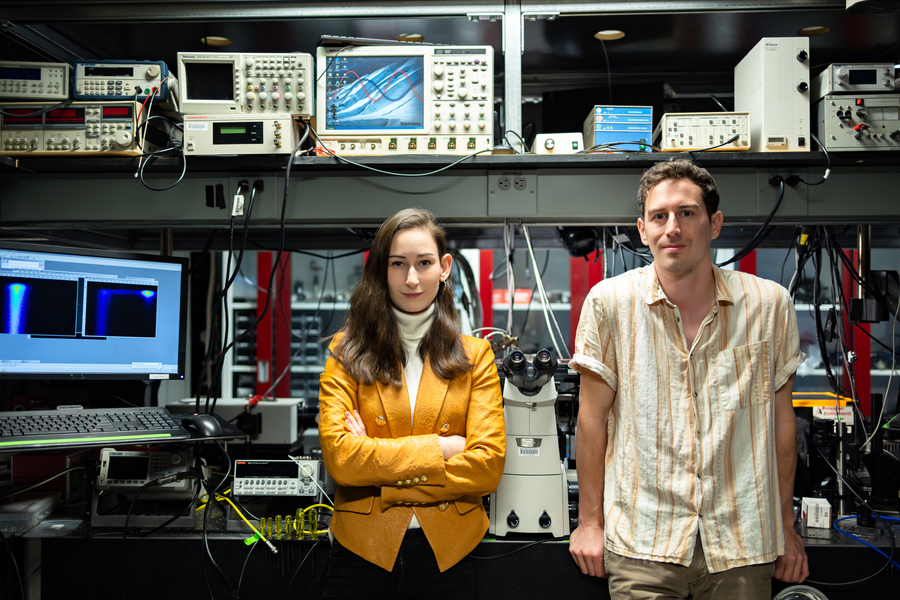 Two MIT researchers stand side by side in a dark electronics laboratory surrounded by oscilloscopes, a microscope, and signal processing equipment, with a fluorescence imaging monitor visible to the left.