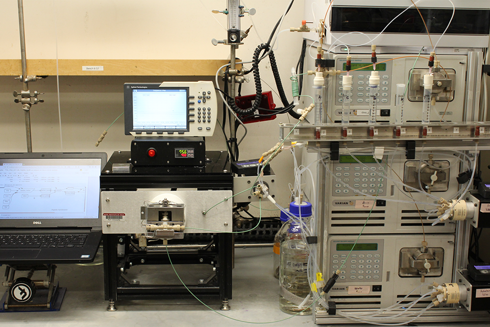 A laboratory benchtop setup featuring a laptop, a compact reactor or instrument, and a rack of chromatography equipment connected by tubing.