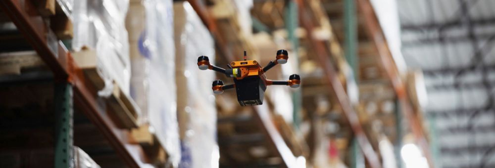 Small drone flying through a warehouse aisle lined with tall shelves stocked with goods.