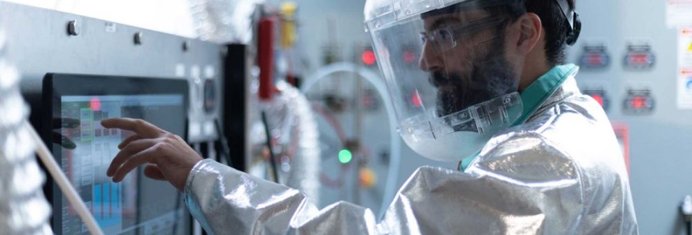 Scientist wearing protective gear and face shield operates a touchscreen panel in a high-tech laboratory.
