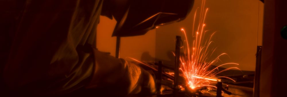 A person wearing protective gear welds metal, with bright orange sparks flying in a dimly lit setting.