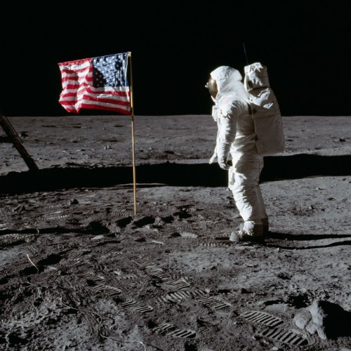 Astronaut in a spacesuit stands on the Moon next to the American flag, with footprints visible on the lunar surface.