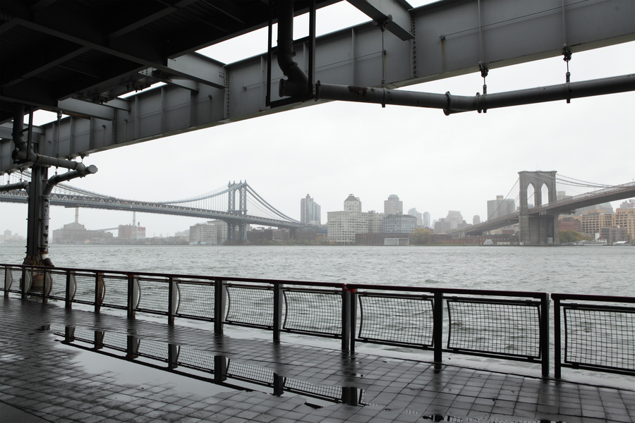 View from a covered pier of the East River on a gray rainy day, with the Manhattan Bridge and Brooklyn Bridge visible through the mist.
