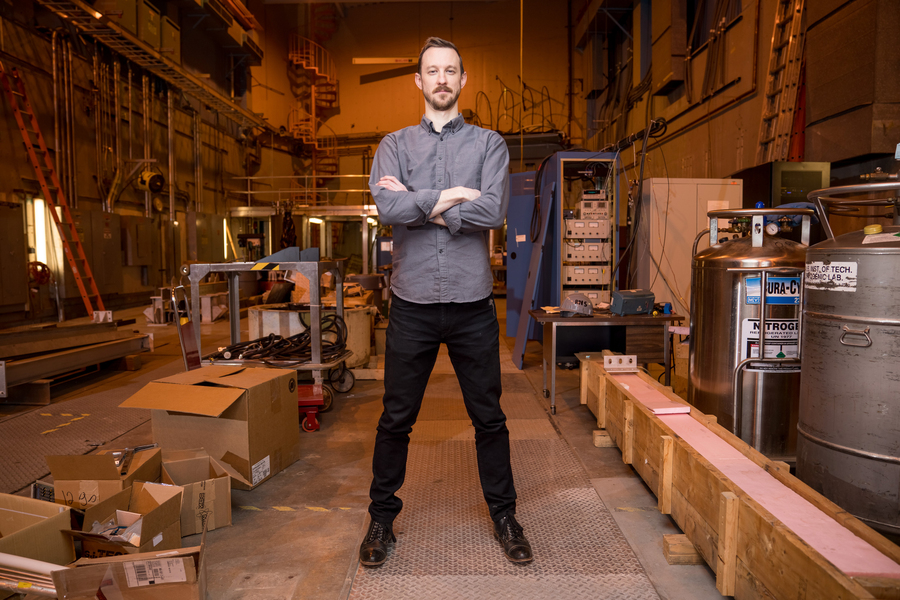 A man with arms crossed stands in a large industrial lab space filled with equipment, cardboard boxes, and a liquid nitrogen tank.