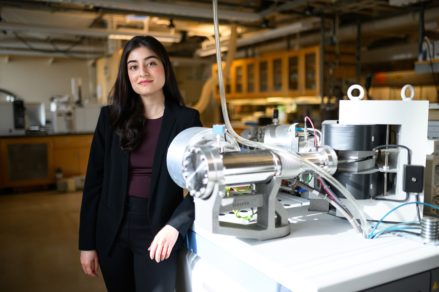A woman in a black blazer stands beside a polished metal scientific instrument on a lab bench, with shelving and lab equipment visible behind her.