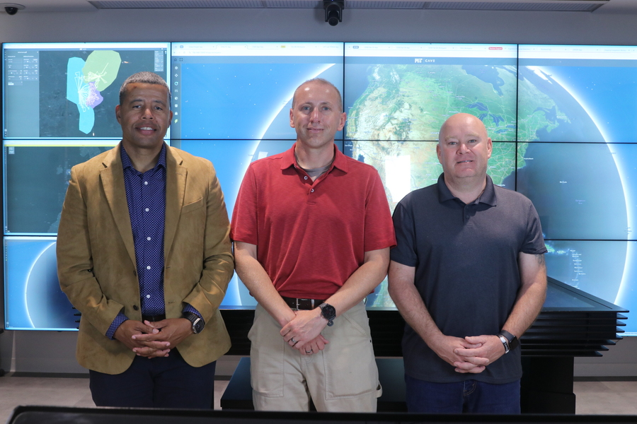 Three MIT researchers stand side by side in front of a multi-screen video wall displaying satellite imagery and a globe visualization in the MIT CAVE facility.