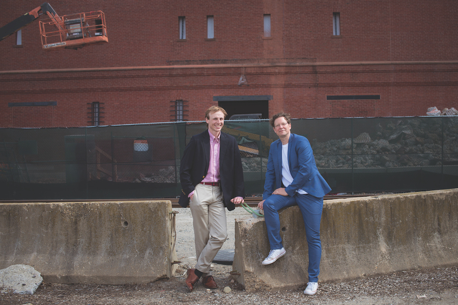 Two MIT researchers pose casually against a concrete barrier at an urban construction site, with a red brick building and scissor lift visible behind them, for a story on urban carbon emissions research.