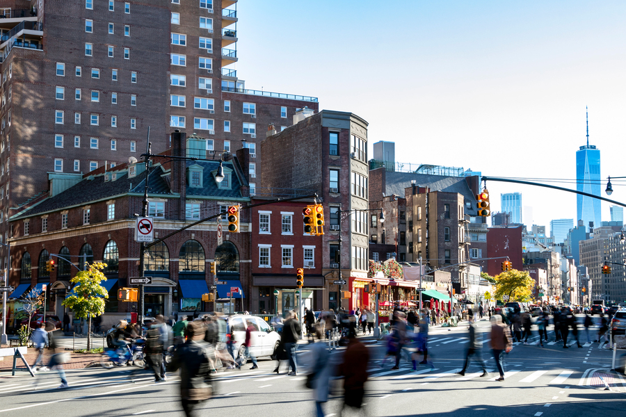 A long-exposure street scene in lower Manhattan with motion-blurred pedestrians crossing a busy intersection, red brick buildings, and One World Trade Center visible in the background.