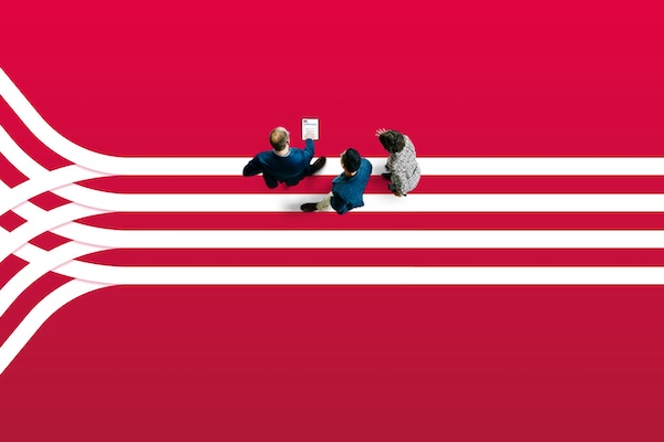 Three people stand on converging white lines against a vibrant red background. One person holds a tablet.