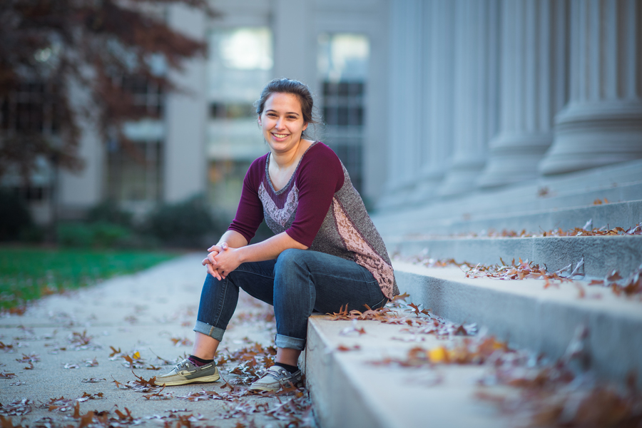 A smiling young woman sits on the stone steps of a columned academic building surrounded by fallen autumn leaves
