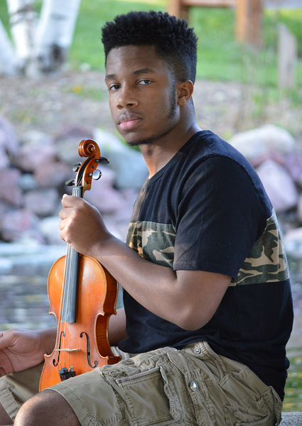 A young man sits outdoors holding a violin, with a natural garden backdrop of birch trees and rocks.