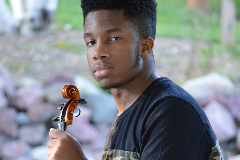 A young man sits outdoors holding a violin, with a natural garden backdrop of birch trees and rocks.